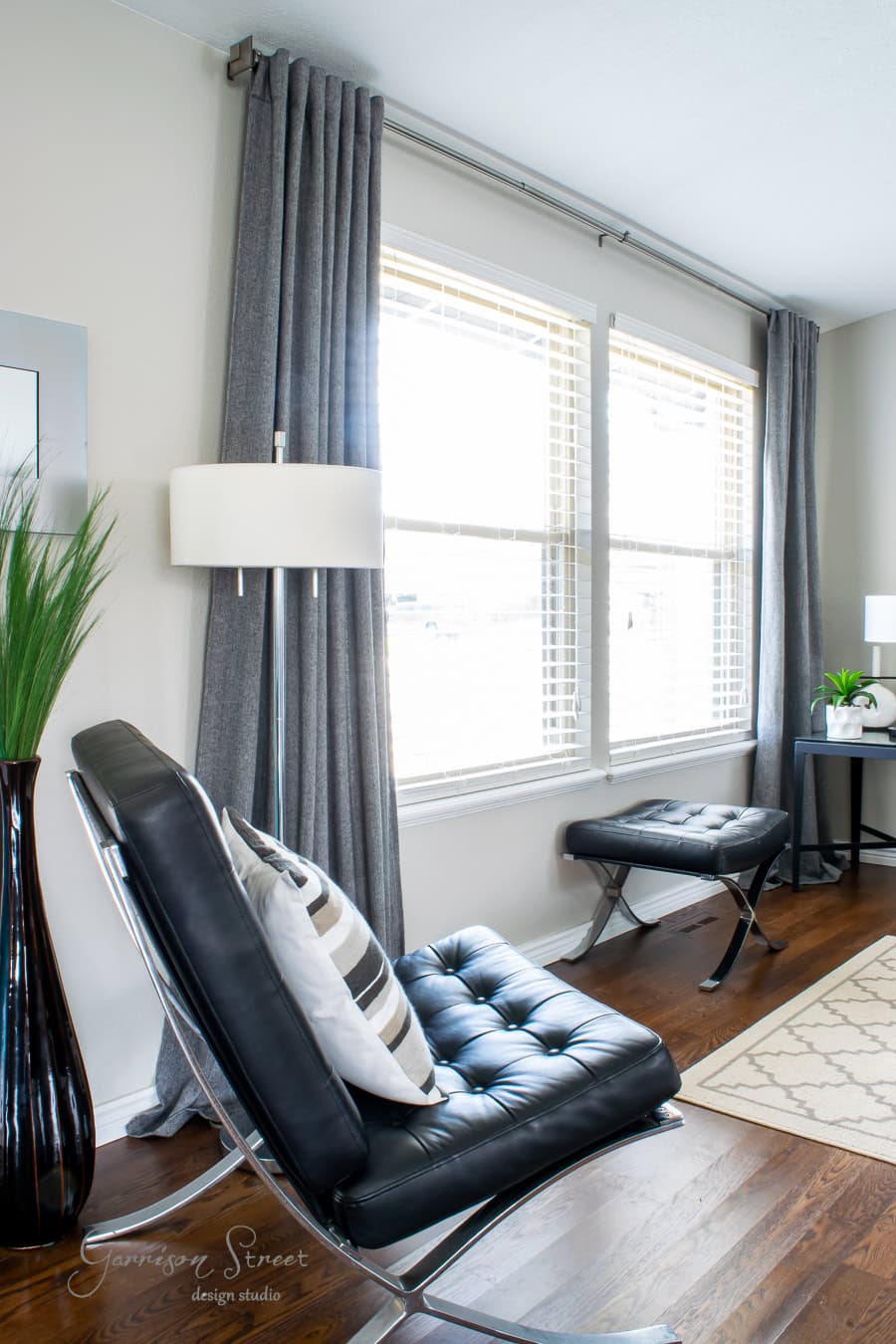 A modern living room showcases a sleek black leather chair and ottoman adorned with a striped pillow, complemented by elegantly hung gray curtains. A floor lamp and a small table with a vibrant plant add charm, while the best feature is the natural light streaming through the large window.