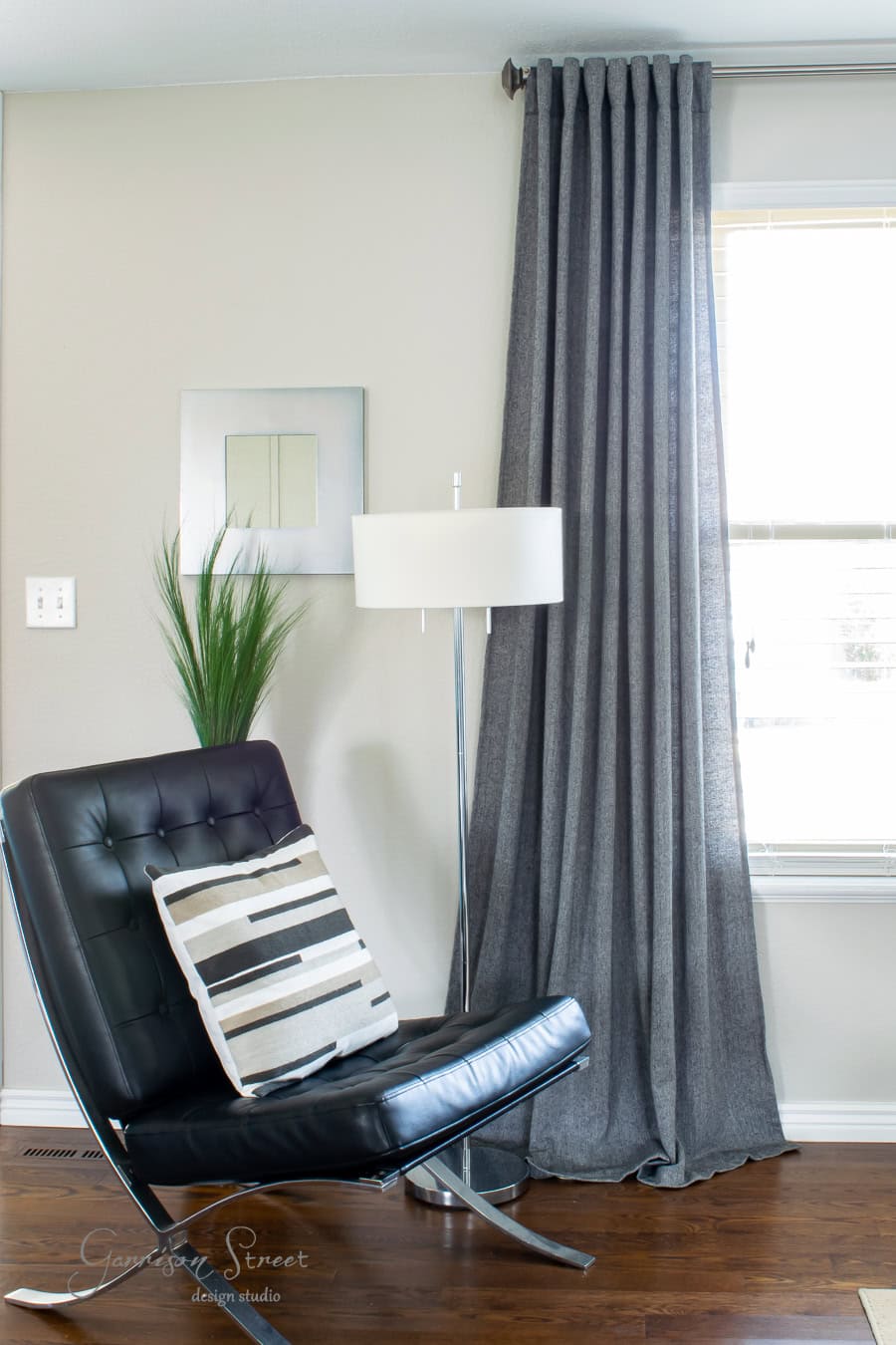 Modern living room corner with a black leather chair, a geometric striped pillow, and the best tall floor lamp illuminating the space. Gray curtains hang gracefully beside a small wall mirror and a green plant. Natural light floods in through the window, brightening every detail.