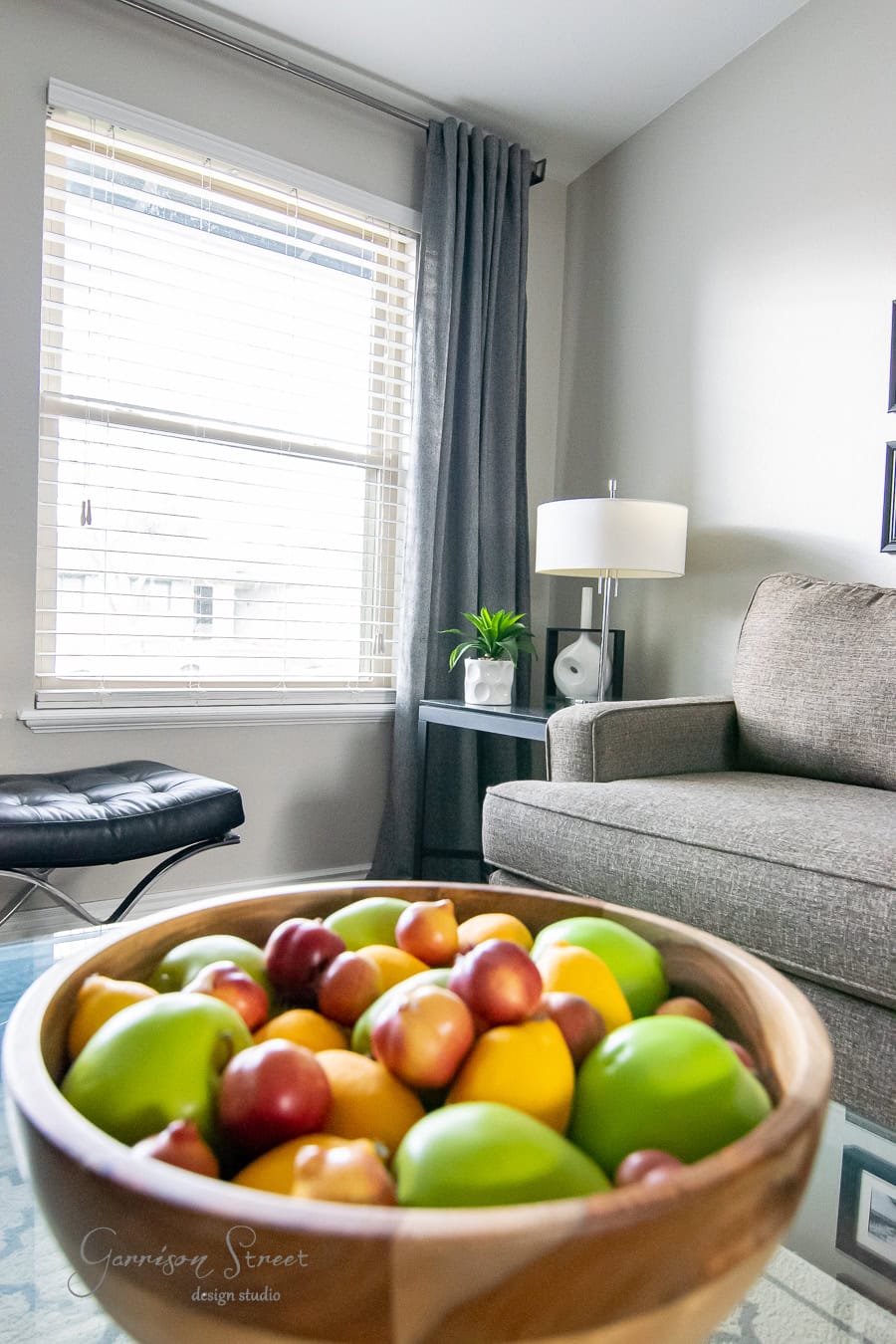 A modern living room with the best gray curtains hanging beside a large window, complemented by a gray sofa. A wooden bowl filled with colorful artificial fruits sits on the glass coffee table, and a small plant rests on a side table next to a lamp.