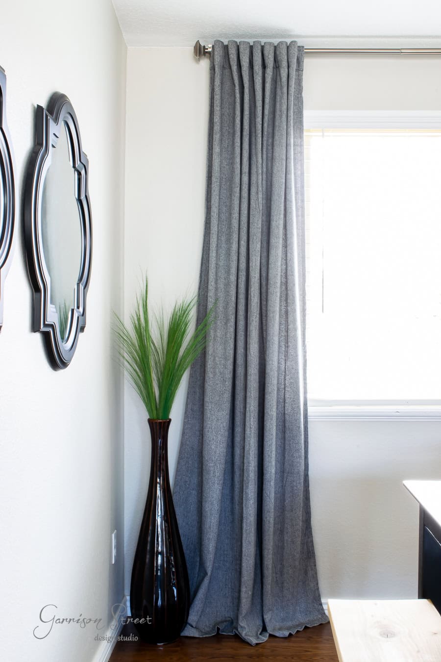 A window with long, gray curtains is partially visible. Next to it stands a tall, dark vase with decorative grass. Adjacent to this, two of the best ornate mirrors hang on a white wall, reflecting parts of the room.