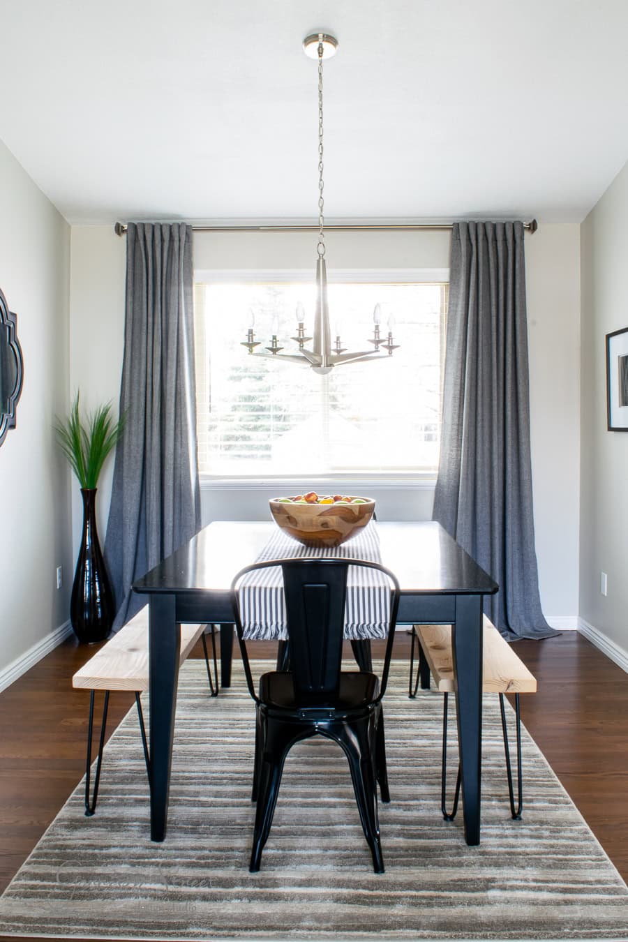 A modern dining room features a black table with a striped runner, flanked by wooden benches and a black metal chair. A bowl of fruit sits in the center. The best gray curtains frame a large window, while a chandelier hangs from the ceiling. A nearby potted plant adds a touch of nature.