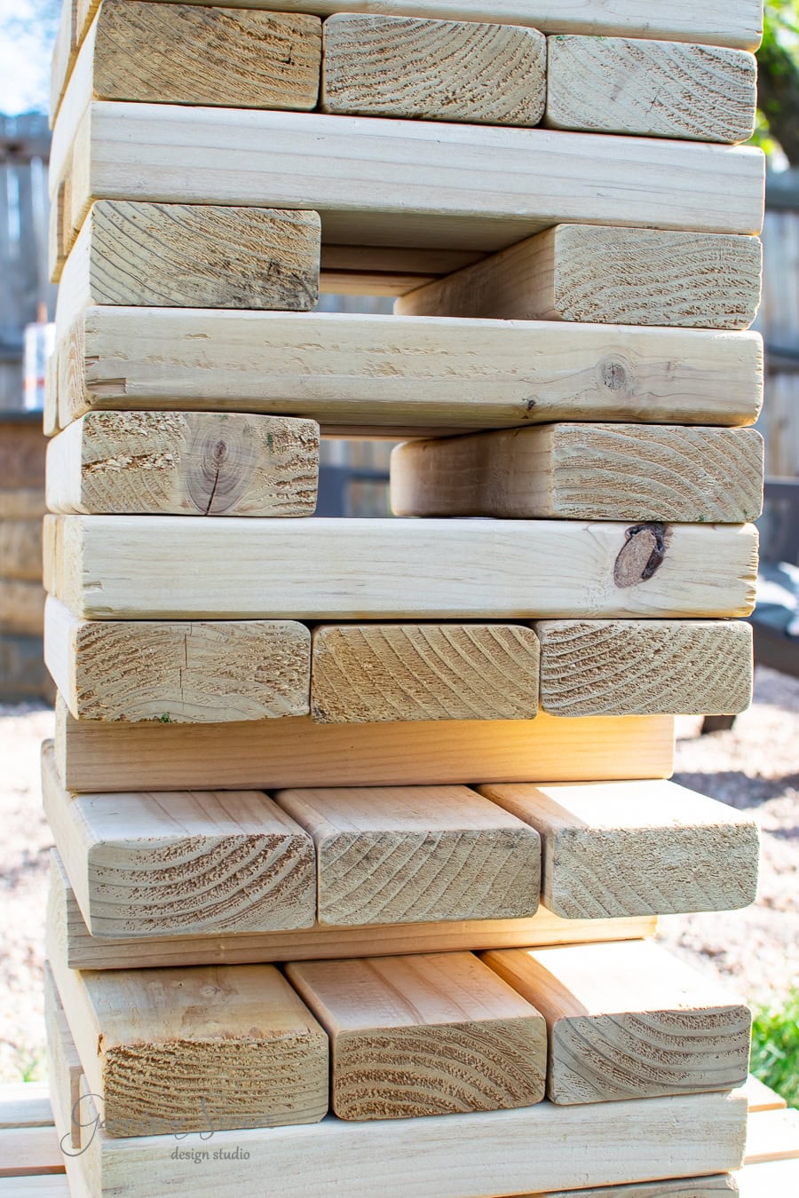Close-up of a DIY Jenga tower made of large wooden blocks, precariously stacked in the yard on a sunny day. One block is partially removed, creating a gap in the structure.