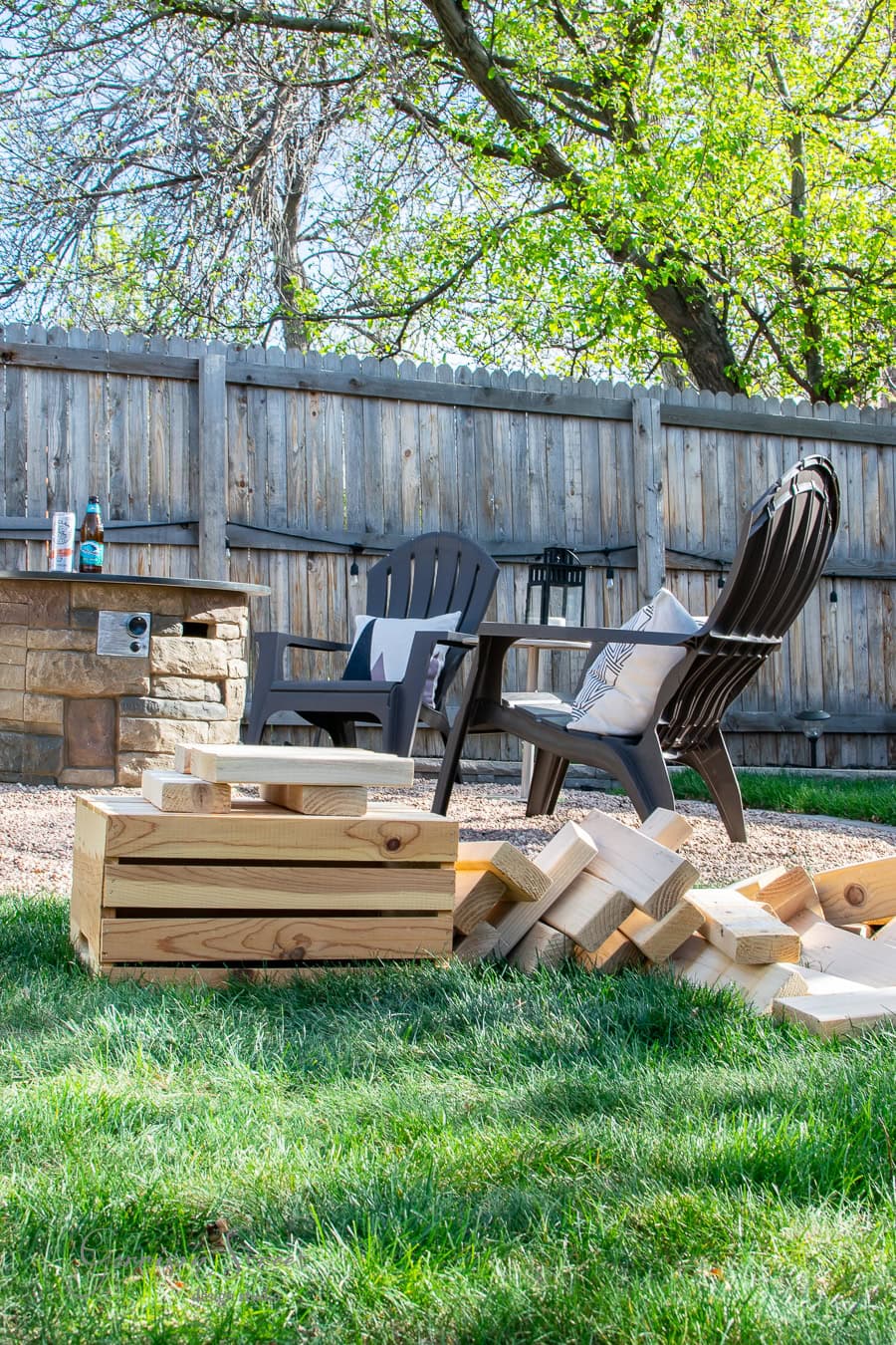 Outdoor DIY area with two plastic chairs by a stone fire pit in a grassy yard. A wooden crate filled with large Jenga blocks awaits on the grass. A wood fence and trees provide a tranquil backdrop, while a cushion and blanket add comfort to one chair.