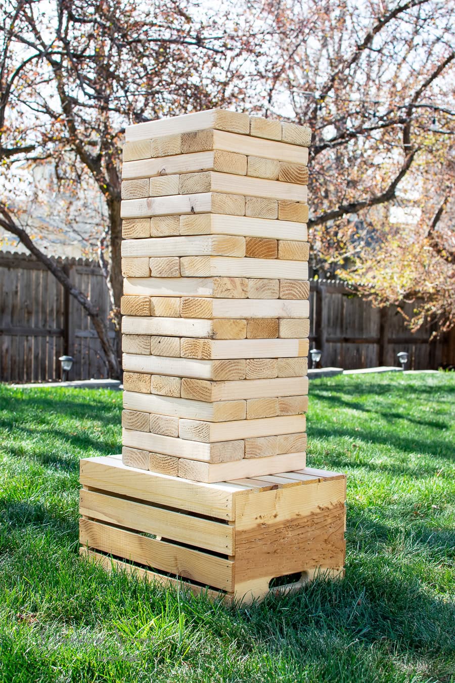 A large DIY Jenga tower stands proudly on the grassy lawn, with trees and a wooden fence in the background. Made of light-colored wood, the blocks are stacked in an alternating pattern on a sturdy wooden crate, perfect for outdoor fun.