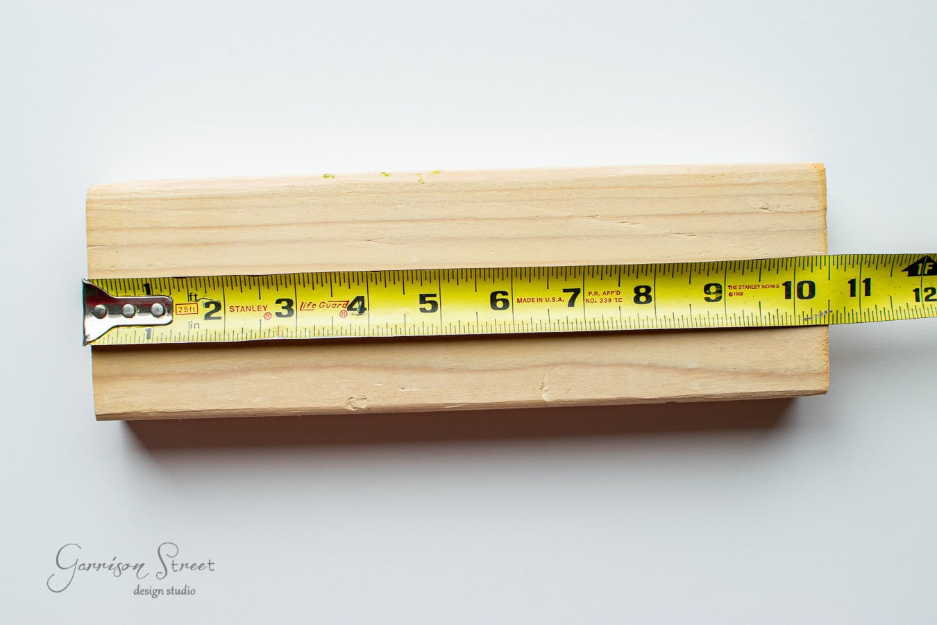A wooden board, perfect for a DIY project like yard Jenga, showcases a yellow measuring tape stretched across it, marking measurements up to 9 inches. Part of the board extends beyond the tape against a plain white background.