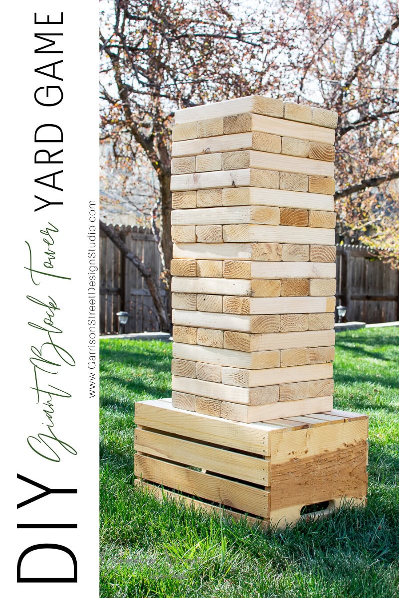 A giant DIY Yard Jenga tower made of wooden pieces stands on a grass lawn. The blocks are stacked on a wooden crate, with trees and a fence visible in the background. The text on the left reads, "DIY Giant Block Tower Yard Game.
