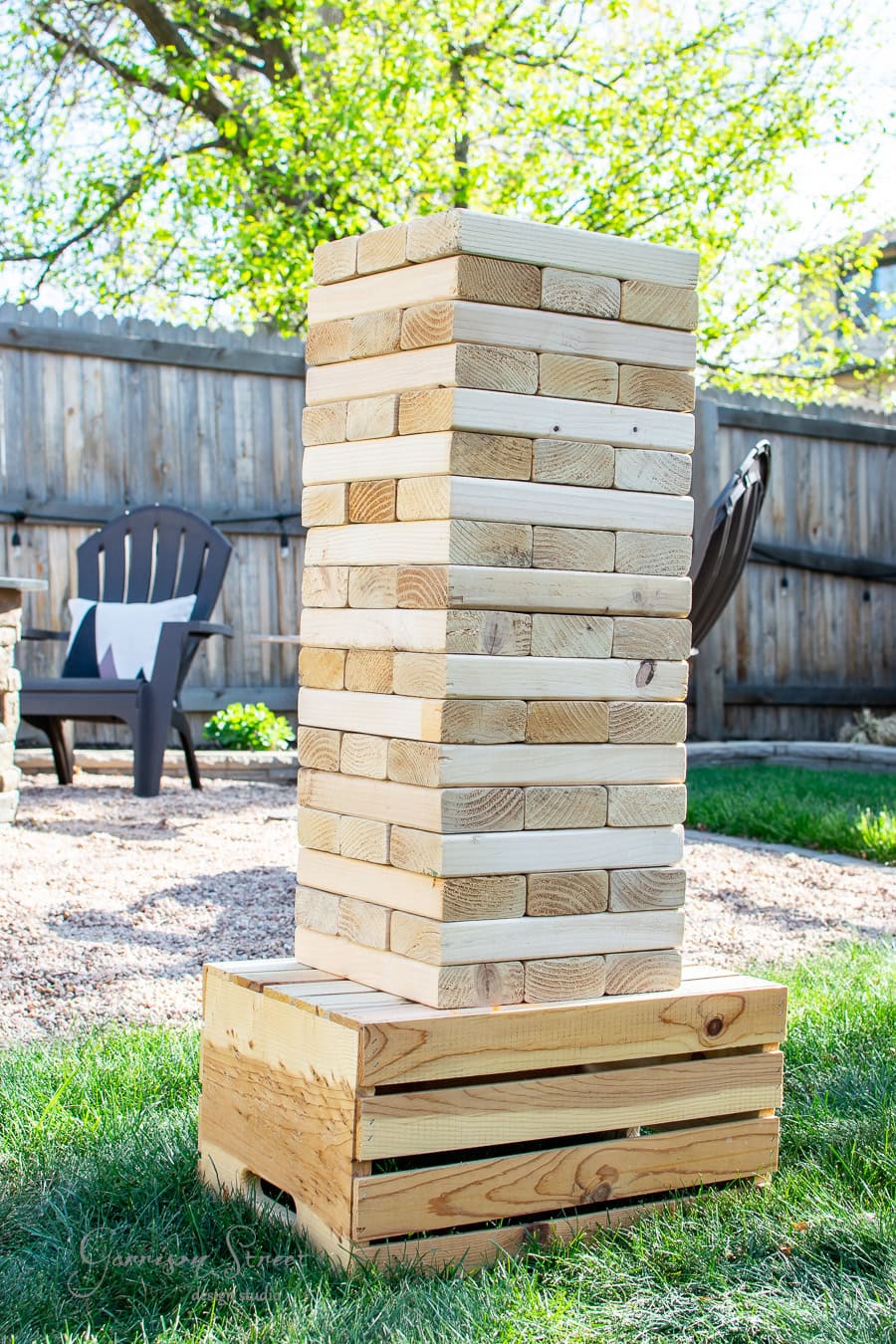 A large DIY outdoor Jenga tower, made of wooden blocks, stands on a grassy surface next to an empty wooden crate. In the background, a fence and black Adirondack chairs rest under a tree on this sunny day, perfect for enjoying your own yard Jenga setup.