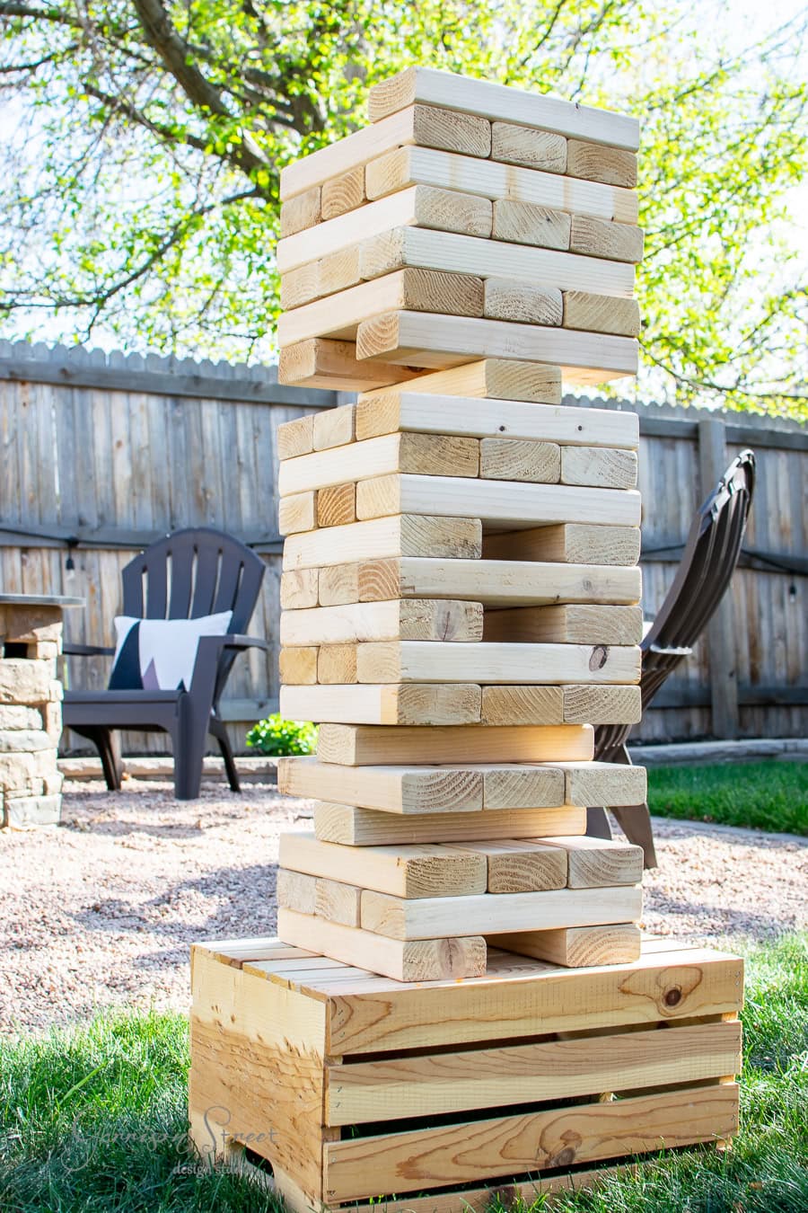 A giant DIY yard Jenga tower made of wooden blocks stands on a wooden crate outdoors. An Adirondack chair and a stone fire pit enhance the charm, with trees and a wooden fence completing the serene backyard setting.