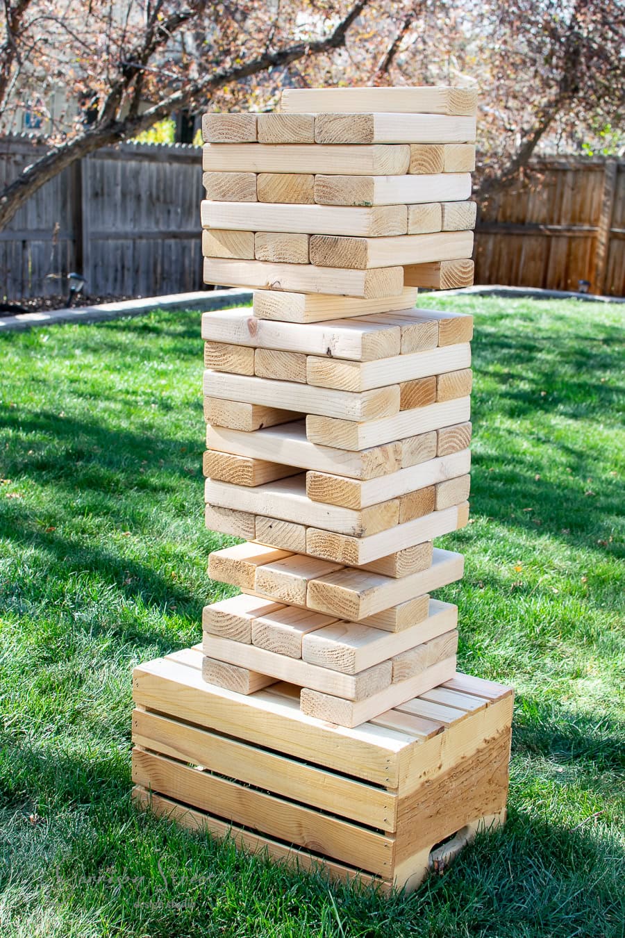 A large DIY Jenga tower made of wooden blocks stands proudly on the grassy lawn, some pieces daringly pulled out. Its base is a rustic wooden crate, and vibrant autumn trees frame the scene, creating a perfect yard attraction.