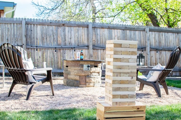 A backyard setting with two Adirondack chairs around a stone fire pit. A large wooden Jenga tower is in the foreground. The area is surrounded by a wooden fence and trees, with bright sunlight casting shadows.