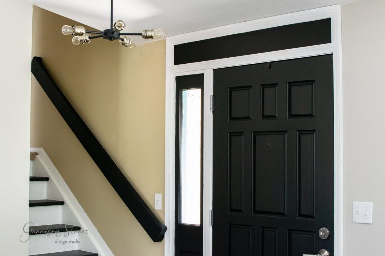 A modern entryway featuring a black front door with a vertical glass panel, a black stair rail, and a contemporary five-bulb chandelier. Neutral walls and a white staircase create a sleek, minimalist design.