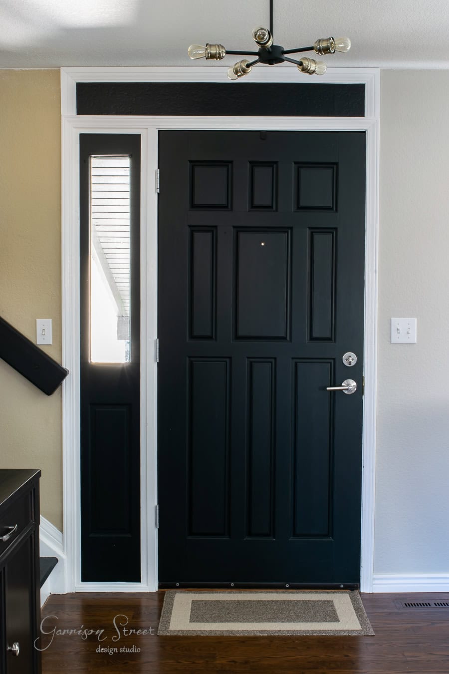 A modern interior with a taller black front door featuring a window panel to the left. A rectangular doormat lies in front, while a ceiling light fixture elegantly hangs above. The walls are beige, and the wooden flooring adds warmth and style to the space.