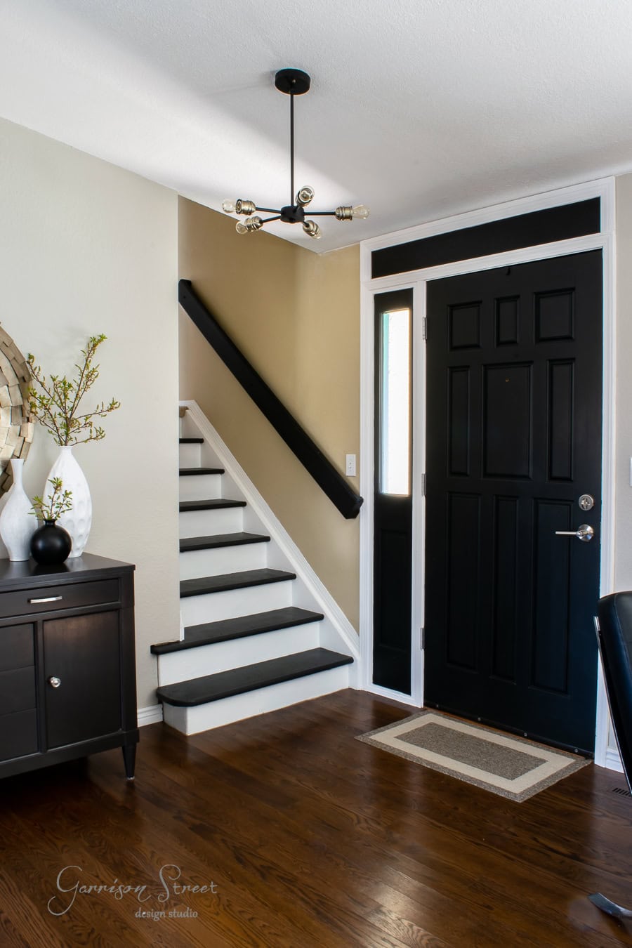 A modern entryway showcases a taller black front door with a glass panel, accompanied by a small stairway featuring black railing and dark wood flooring. Beside the stairs, a sleek black cabinet adorned with decorative items complements the contemporary ceiling light overhead.