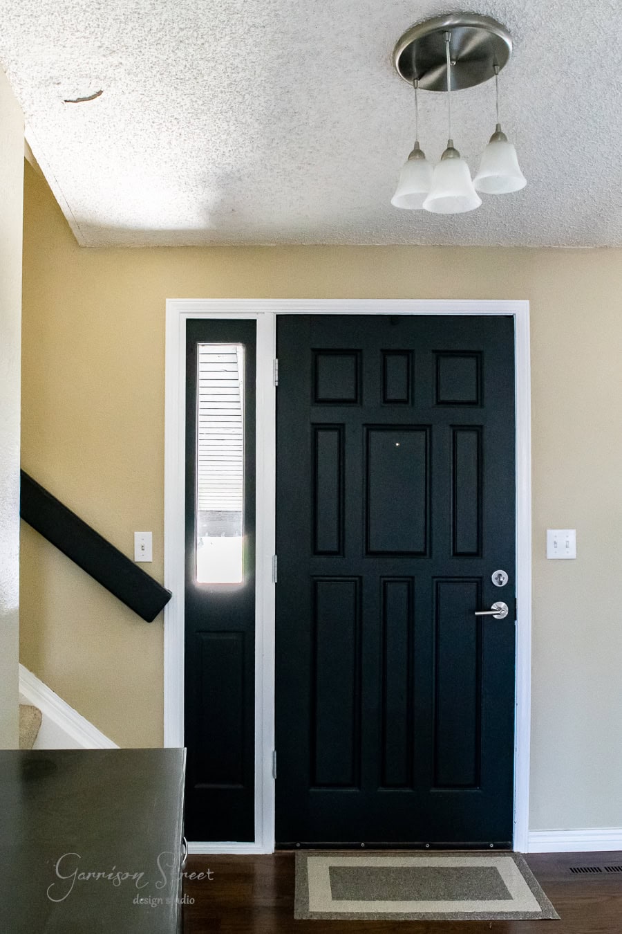 The image depicts a simple entryway with a taller black front door featuring a narrow window on its side. The beige walls complement the small staircase with a black handrail on the left. Above, a ceiling light with three white glass shades illuminates the space.