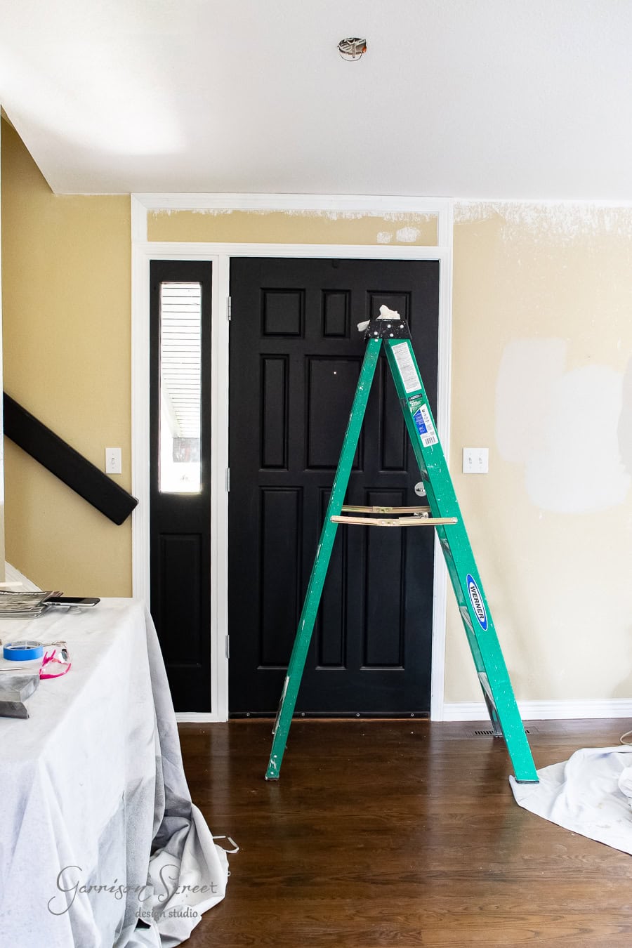 A room under renovation features a taller, black front door. A green ladder stands before it, with paint patches adorning the beige walls. Nearby, a covered table holds painting supplies on the wooden floor.