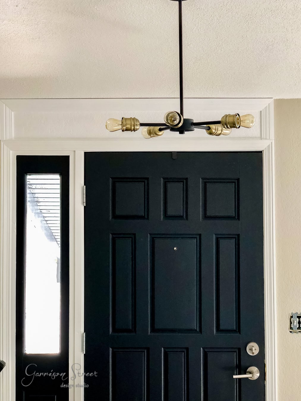 A modern light fixture with exposed bulbs hangs above the taller black front door, featuring a silver handle and a peephole. Encased in a white frame, it is complemented by a small side window panel on the left, while the interior wall boasts a warm beige hue.
