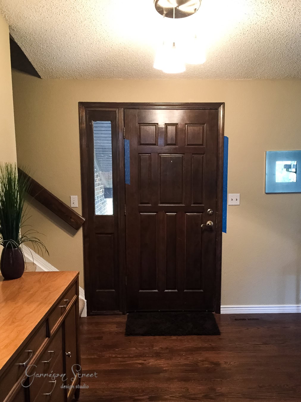 The entrance hallway features a taller dark wooden front door flanked by a narrow glass pane. The hardwood floor complements the cabinet adorned with decorative greenery on the left, while a small illuminated artwork graces the right wall. A ceiling light fixture brightens the space.