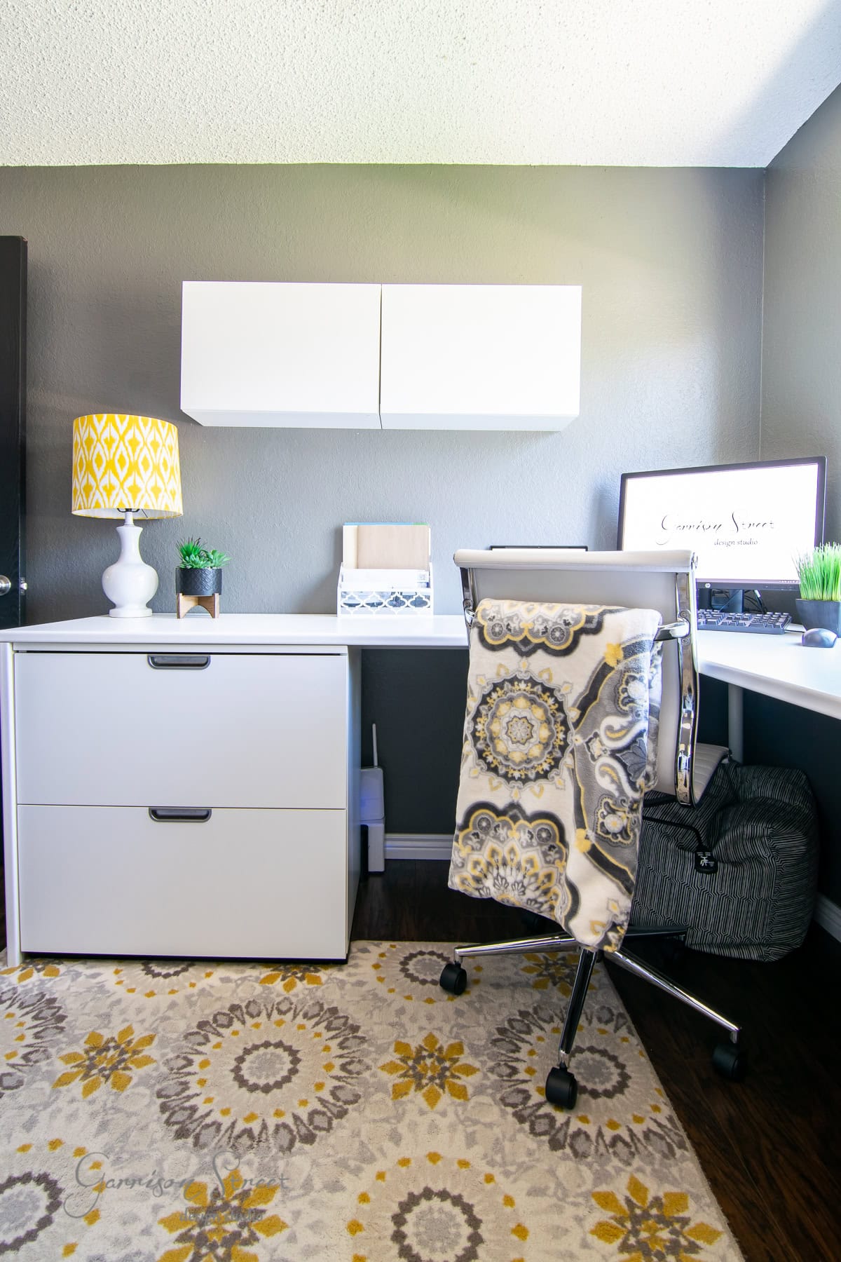 A modern home office featuring sleek white IKEA furniture, including a desk and filing cabinet. A patterned chair, geometric lamp, and wall cabinets enhance the space, while a colorful rug with circular patterns brightens the dark wood floor.