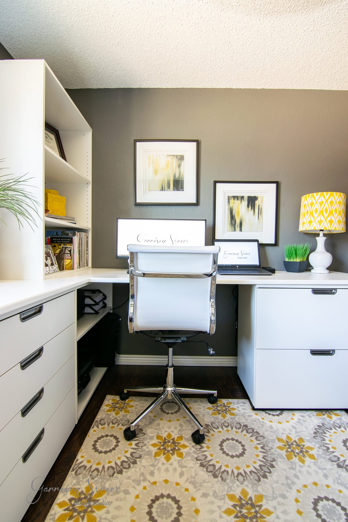 Modern home office featuring an IKEA white desk, swivel chair, and wall art above dual monitors. The room includes a yellow-patterned lamp, bookshelves with decor, and a floral rug on a dark floor. The walls are painted a soft gray, creating a stylish and functional workspace.