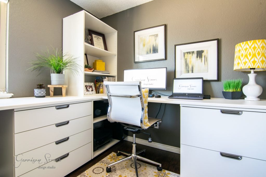 Home A modern home office with a white desk, swivel chair, and open shelving. Two computer screens are on the desk. Framed abstract art hangs on the gray walls, and a yellow patterned lamp and potted plants add color. A patterned rug is on the floor.