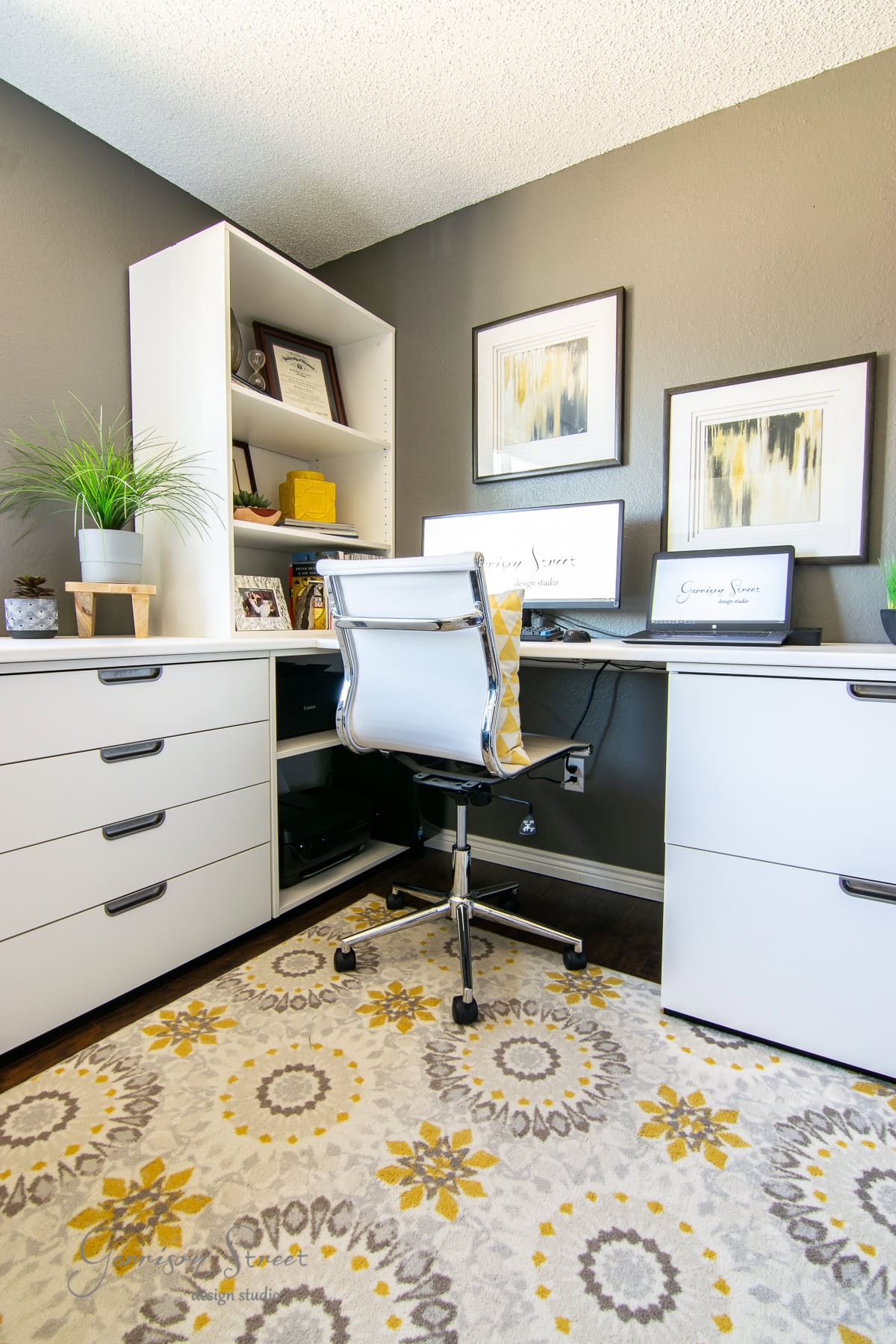 A modern IKEA-inspired home office with a white swivel chair, two monitors on a sleek white desk, and minimalist shelves showcasing books and decor. The walls feature framed art, while a yellow and gray patterned rug adds warmth to the dark floor.