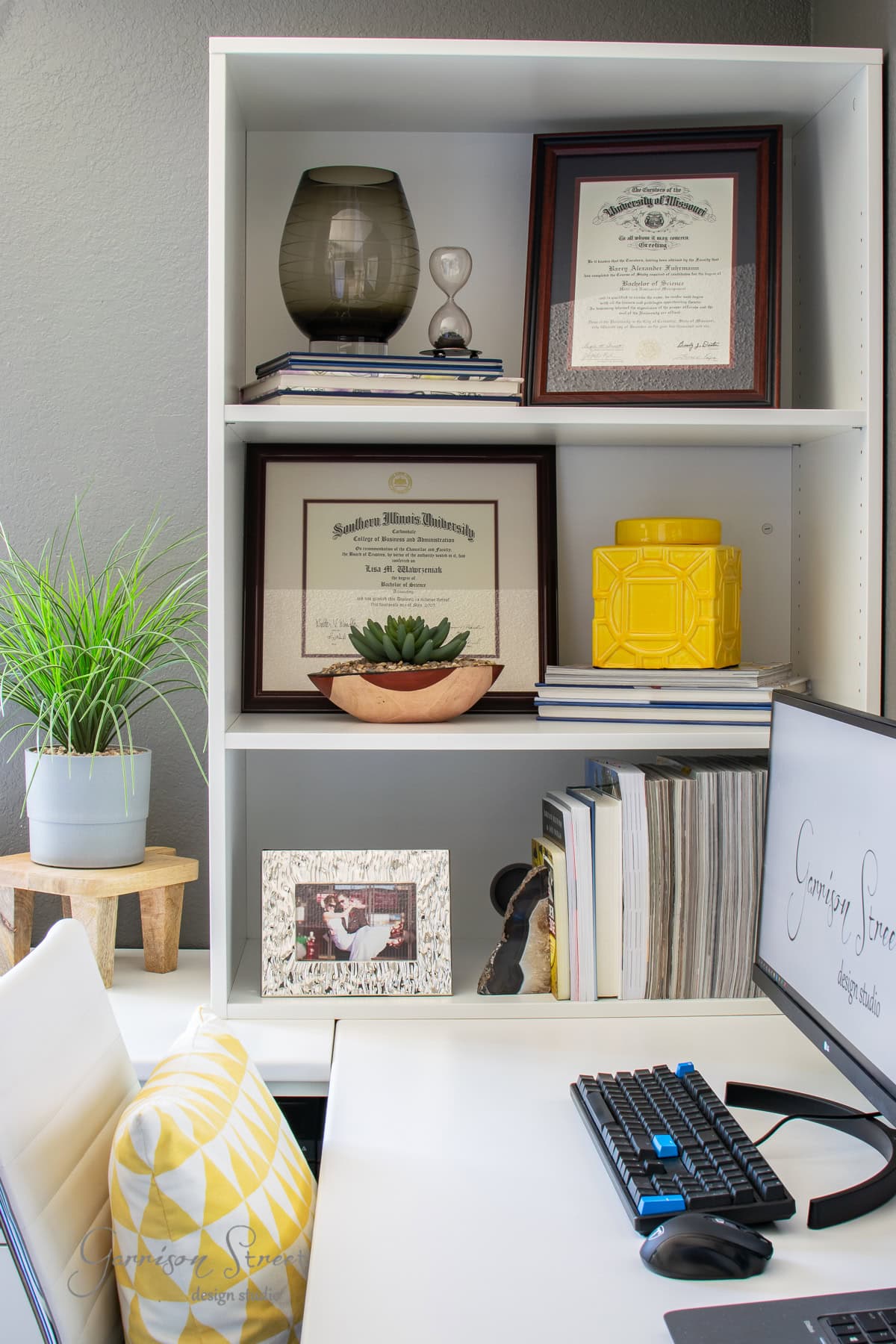 A tidy home office showcases an Ikea white desk with a keyboard, adorned with a patterned yellow pillow. Shelving above holds diplomas, books, a yellow jar, a potted plant, and framed photos. A vase and other decor items add a personal touch.