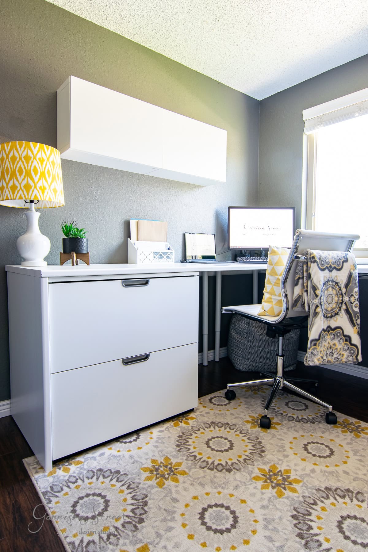 A modern IKEA home office features a white desk and filing cabinet, a patterned rug, and a yellow lamp. A white shelf hangs above the desk. The office chair boasts a colorful cushion, and sunlight streams through the window on the right.