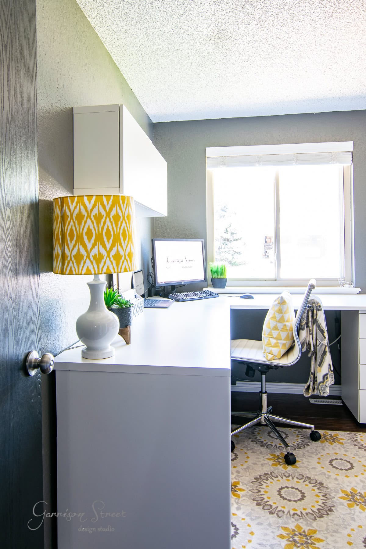 A modern home office with a sleek IKEA white desk and chair facing a window. The desk features a computer, a lamp with a yellow patterned shade, and small plants. The room's gray walls complement the patterned yellow and gray rug perfectly, creating an inviting workspace.