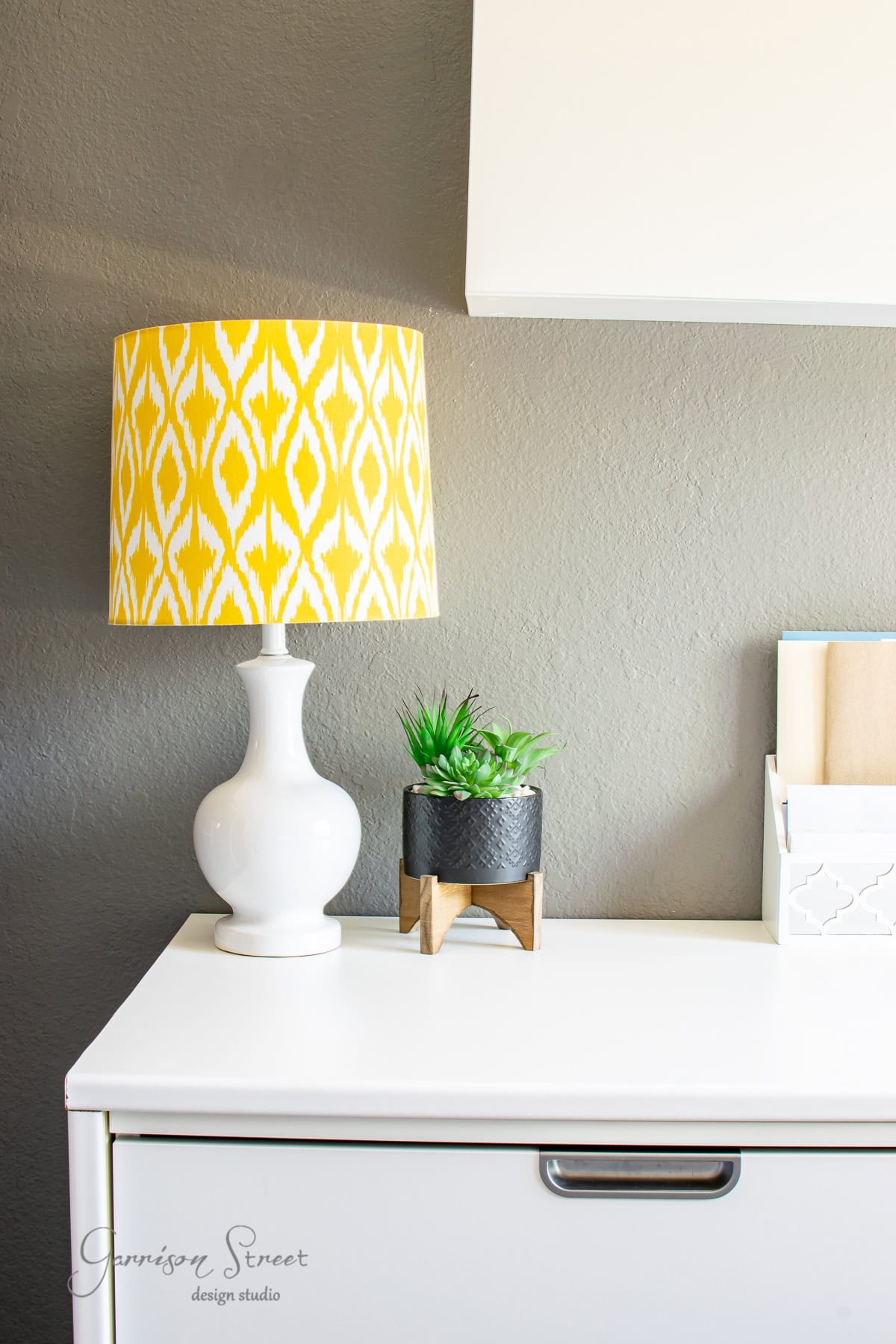The white desk, accented by a yellow patterned lamp and a small potted succulent, creates a cozy IKEA-inspired office vibe. The gray wall backdrop features a sleek white shelf.