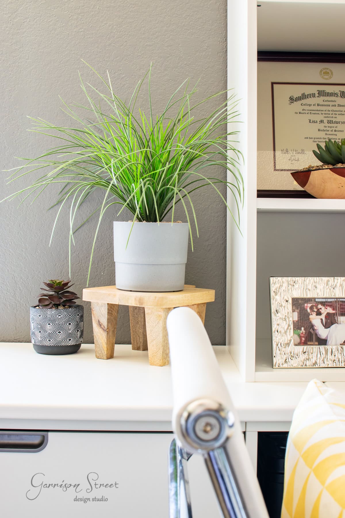 A modern home office setup featuring a sleek white desk with plants evokes a sense of calm. A small succulent in a textured pot and a taller plant in a gray pot on a wooden stand add greenery. A diploma and photo frame rest on an Ikea shelf, while a white chair completes the scene.