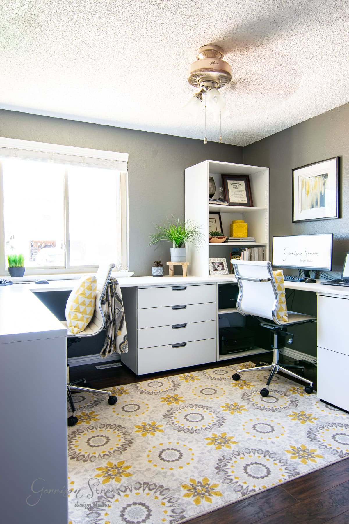 A modern home office with gray walls features white Ikea desks and swivel chairs. A computer screen displays a design. The room has a window, a small potted plant, wall art, and a patterned rug with yellow and gray circles, illuminated by a ceiling fan light.