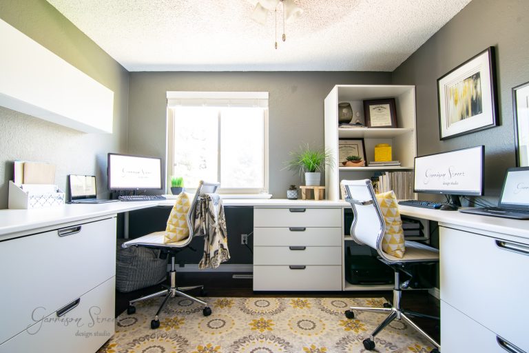 A modern home office features two desks with white chairs, a patterned rug, and a window in the center. Laptops and decorative items are neatly arranged on the desks and shelves, complemented by gray walls and white furniture.