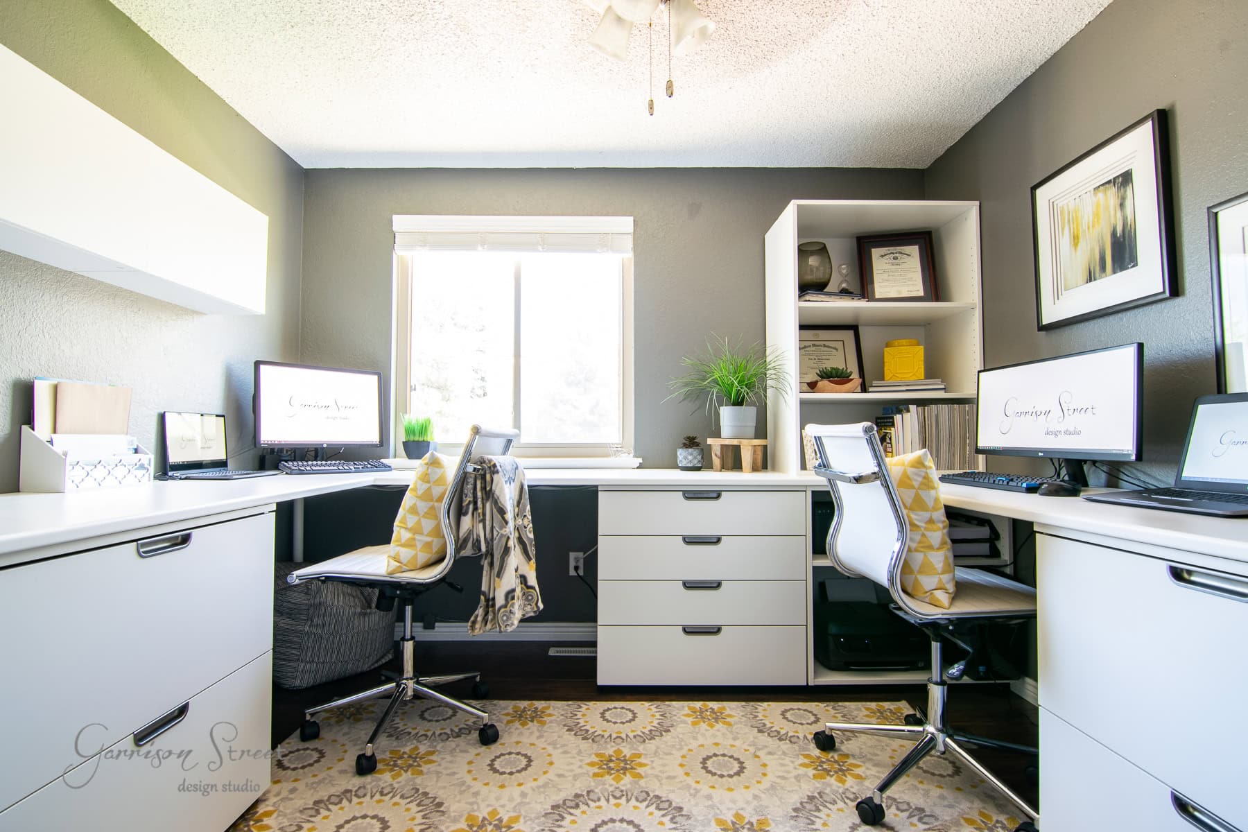 A modern home office features two desks with white chairs, a patterned rug, and a window in the center. Laptops and decorative items are neatly arranged on the desks and shelves, complemented by gray walls and white furniture.