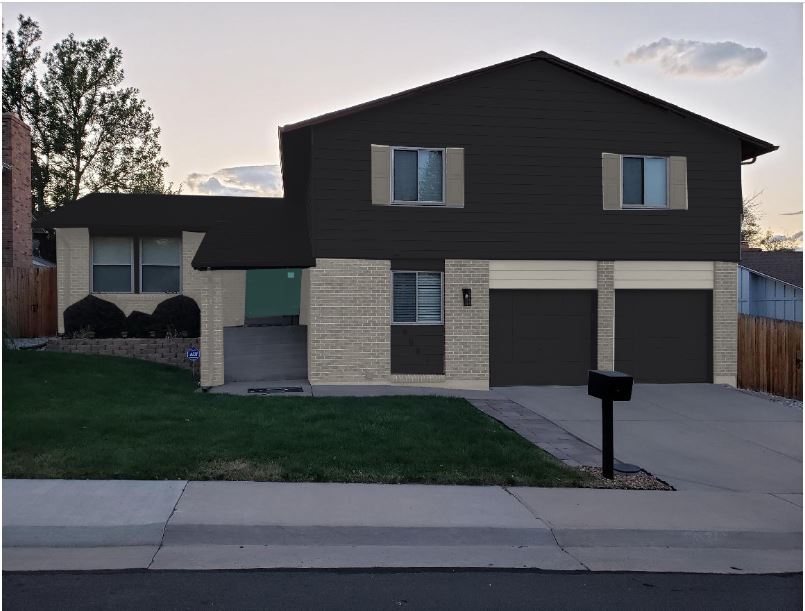 A split-level house with a striking dark brown and beige brick exterior features two sleek black garage doors. It boasts a small front lawn with a sidewalk, mailbox, and verdant trees in the background under a partly cloudy sky.