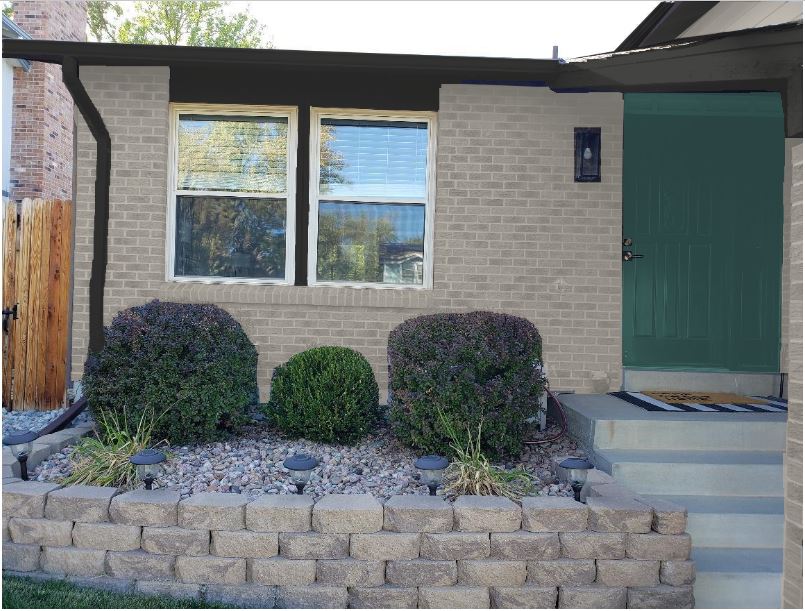 A house entrance with a classic brick facade, two large windows, and a green door. The small garden in front features neatly trimmed bushes and landscaping rocks, bordered by a short stone wall. A black doormat lies at the top of the steps.