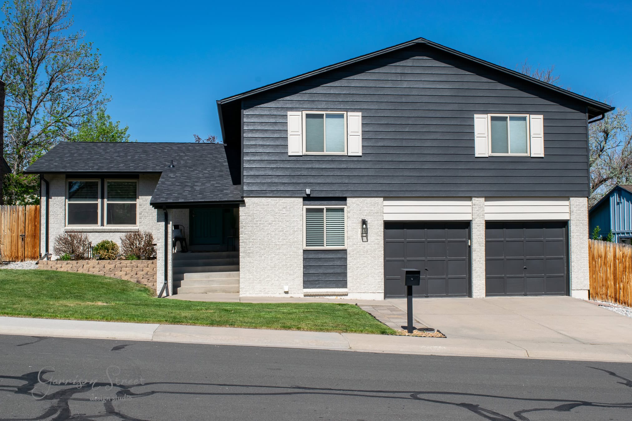 A two-story house boasts a dark gray exterior with white trim and subtle brick accents, complemented by a two-car garage. The front features a small lawn with a few bushes, and a paved driveway leading to the street. Under the clear blue sky, the home presents an elegant facade.