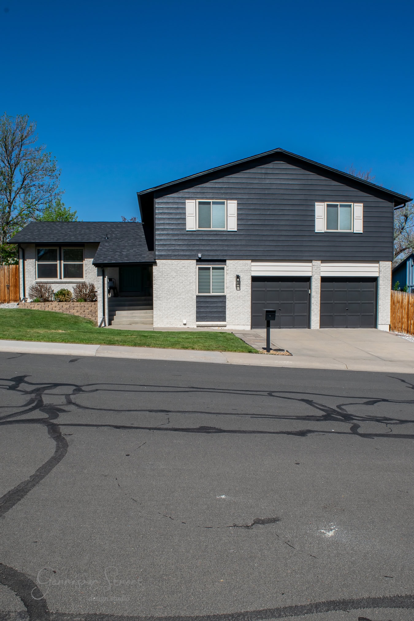 A modern two-story house with black and white exteriors that resemble sleek brushstrokes of paint. It has a double garage, a small front lawn, and a few bushes. The sky is clear and blue, with the house bordered by a sturdy wooden fence.