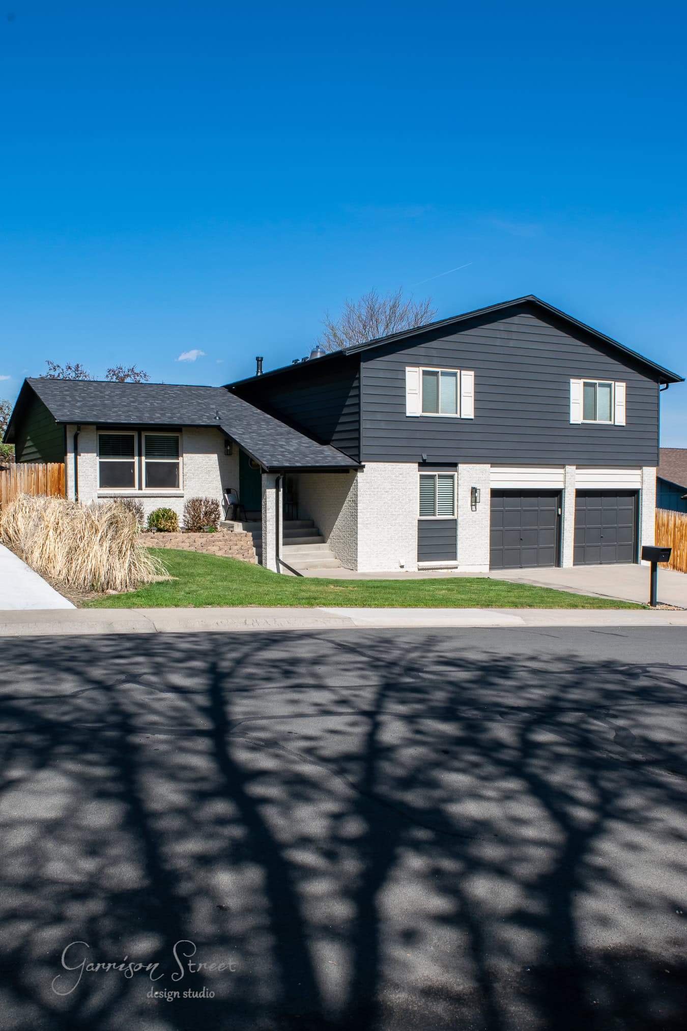 A modern two-story house with a white and dark gray exterior, accented by sleek black details, is surrounded by a lawn and a few shrubs. The driveway leads to a two-car garage, while tree shadows dance on the street under a clear blue sky.