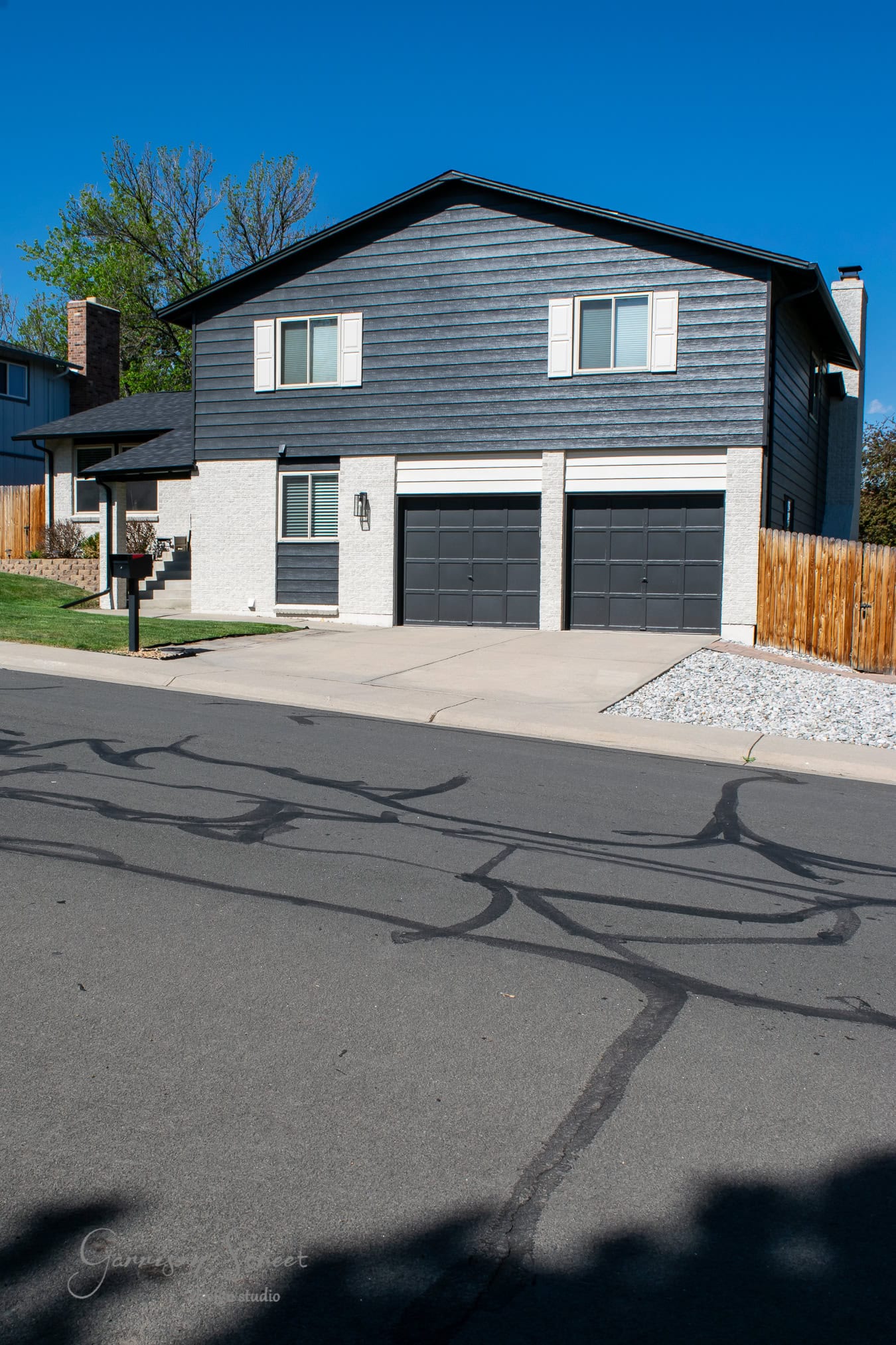 A two-story house with dark siding and white accents features a double garage. The front yard boasts a small lawn, a mailbox, and a paved driveway. A wooden fence lines the right side, contrasting beautifully against the black sky above.
