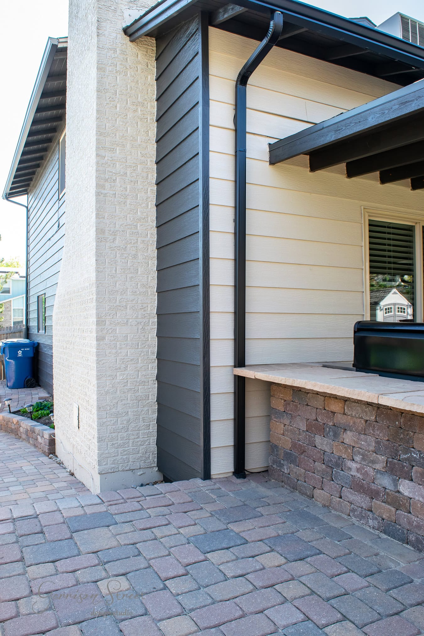 Side view of a house with two different wall textures, one light brick and one modern grey paneling. There's a small patio with stone accents, black trim, and a visible rain gutter. Brick patio flooring extends alongside a path with a recycle bin.