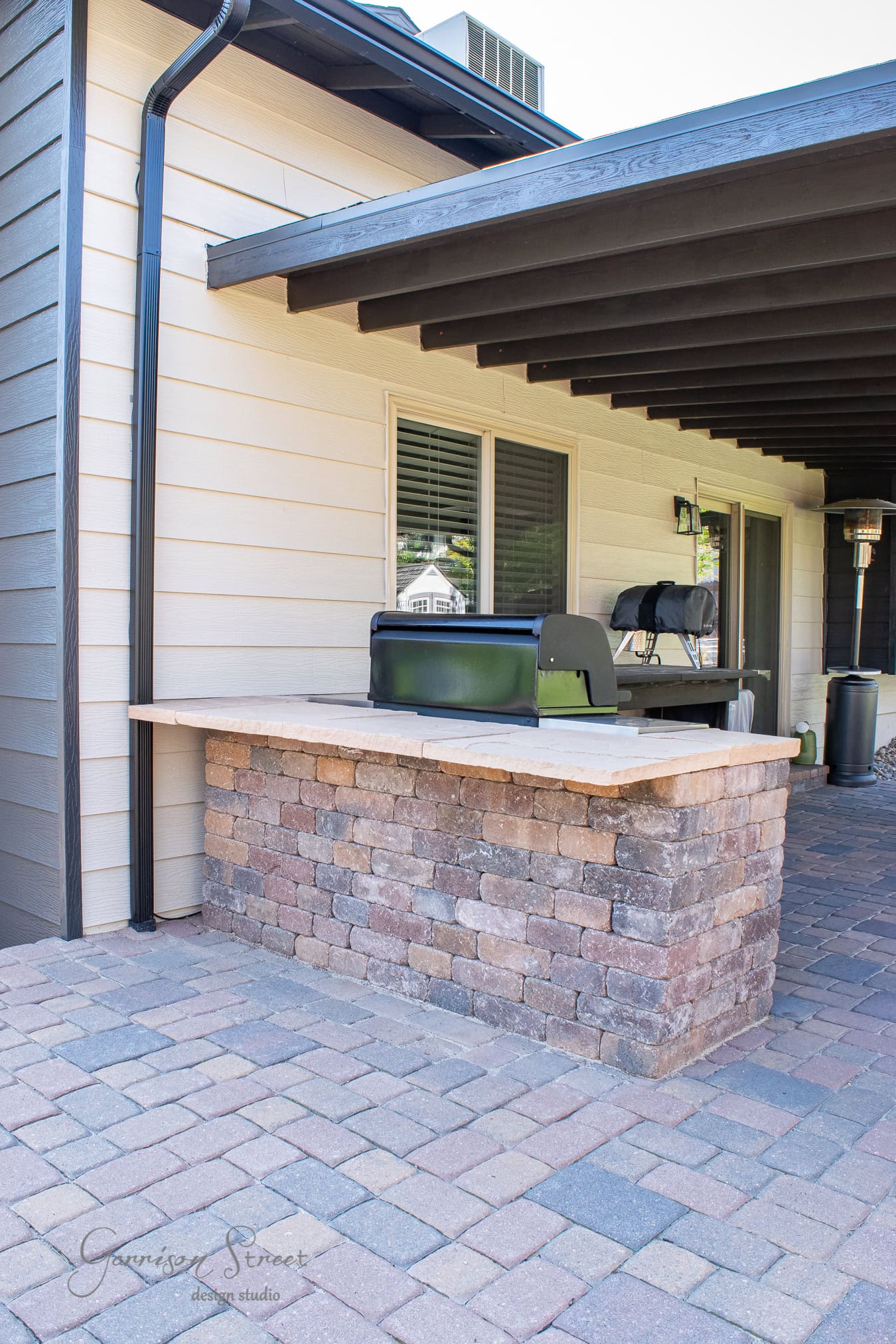 Outdoor barbecue setup under a wooden pergola. The grill is built into a stone countertop, surrounded by a patio with brick paving. The house exterior features light-colored walls with sleek black trim, providing a striking contrast to the inviting space.