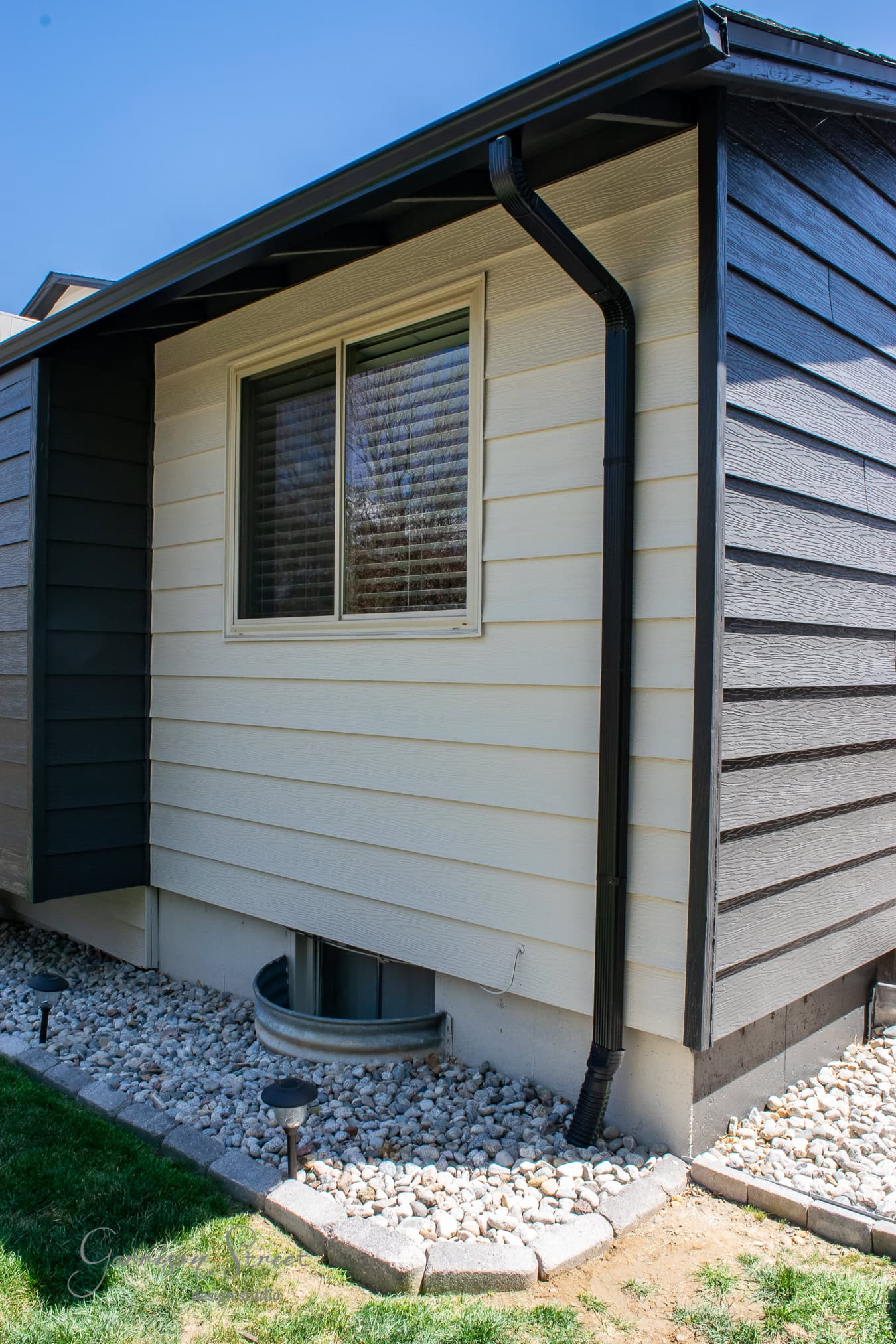 A corner view of the house exterior showcases beige and gray siding with a black gutter. Accentuating the window is a small rock garden, while a well-maintained green lawn completes the scene. A hint of fresh paint enhances the classic appeal.