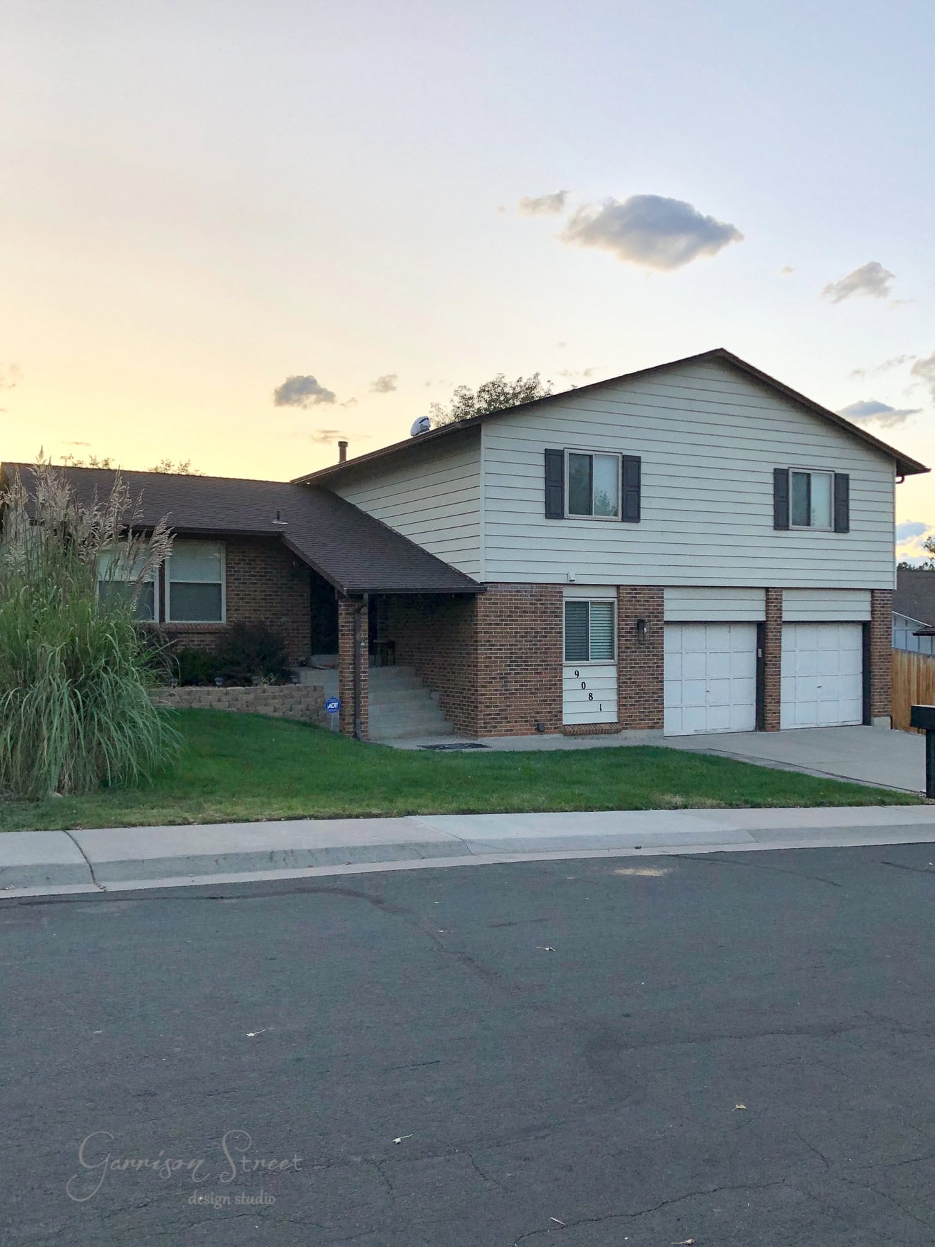 A suburban split-level house with brick and white siding, featuring two black garage doors. The façade, enhanced by a fresh coat of paint, is set against a clear sky with a few clouds. In the foreground, a small lawn dotted with ornamental grass completes the picturesque scene.
