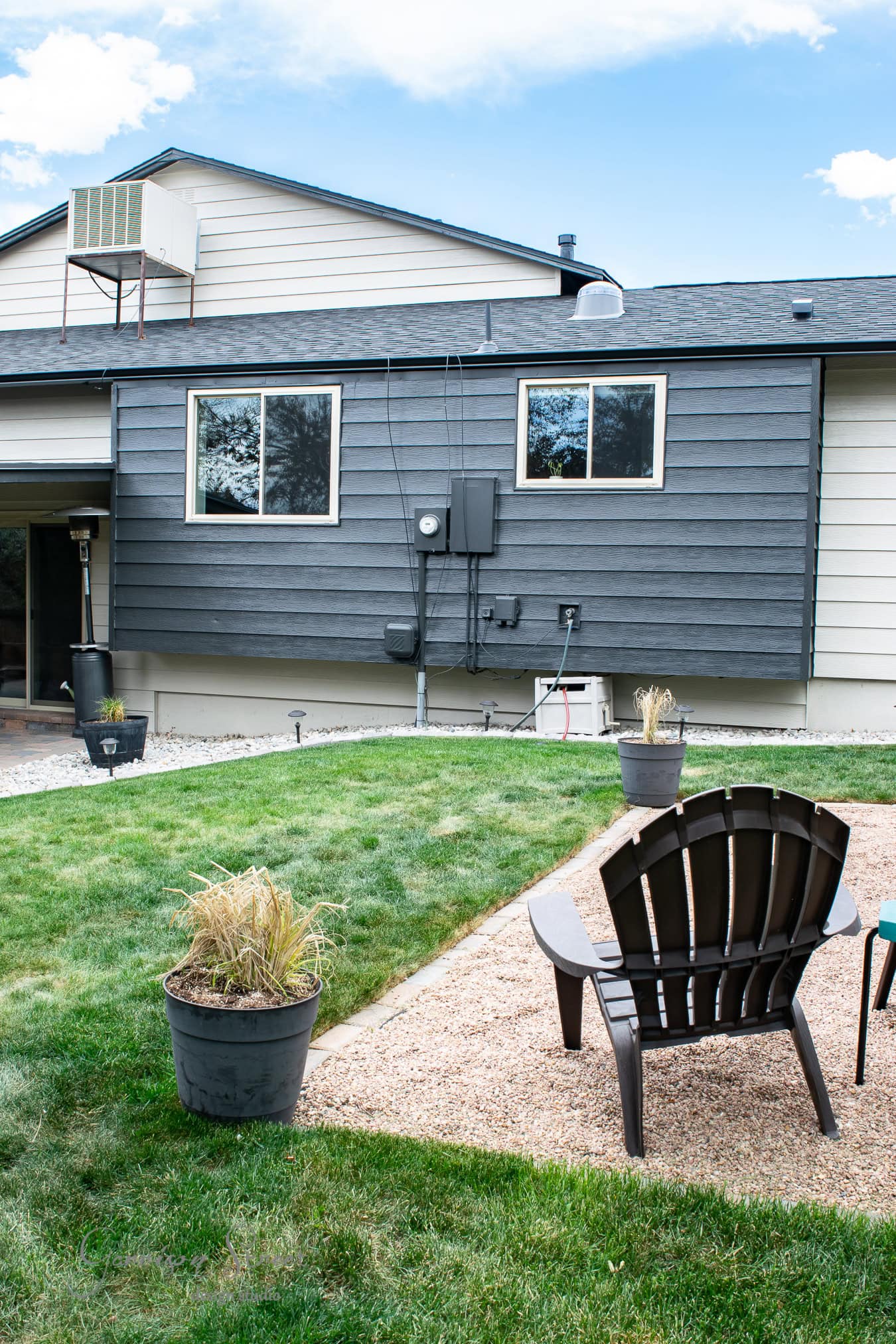 Backyard view of a modern house with dark gray siding and a striking black accent wall. The yard features a well-maintained lawn, a gravel area, and a wooden Adirondack chair. Potted plants are scattered around, and the sky is partly cloudy.