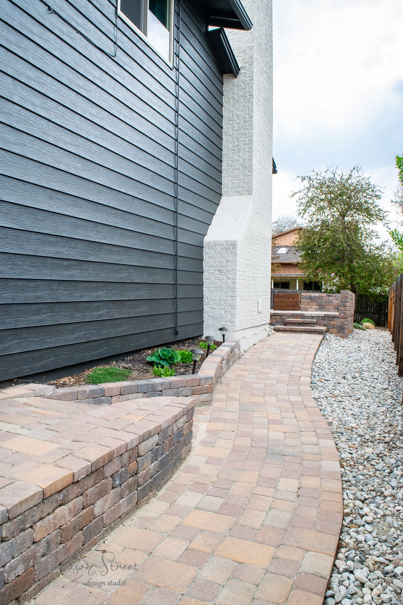 A modern house exterior features a paved brick walkway alongside a sleek gray wall. The path, accented with black stone borders and gravel, leads to a garden area with vibrant greenery and two trees under the cloudy sky.