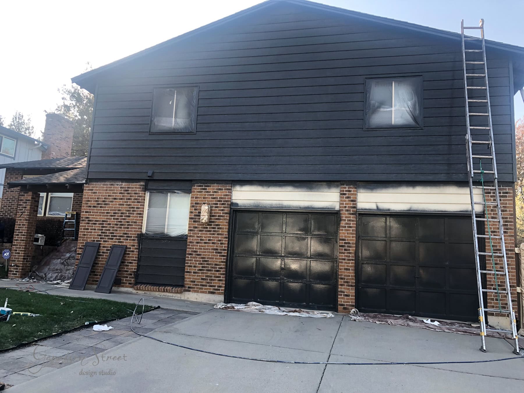 A two-story house with black brick and dark siding is being painted. The lower portion, including three garage doors, is partially covered with plastic sheets. A ladder leans against the right side of the house, and some painting supplies are scattered on the driveway.