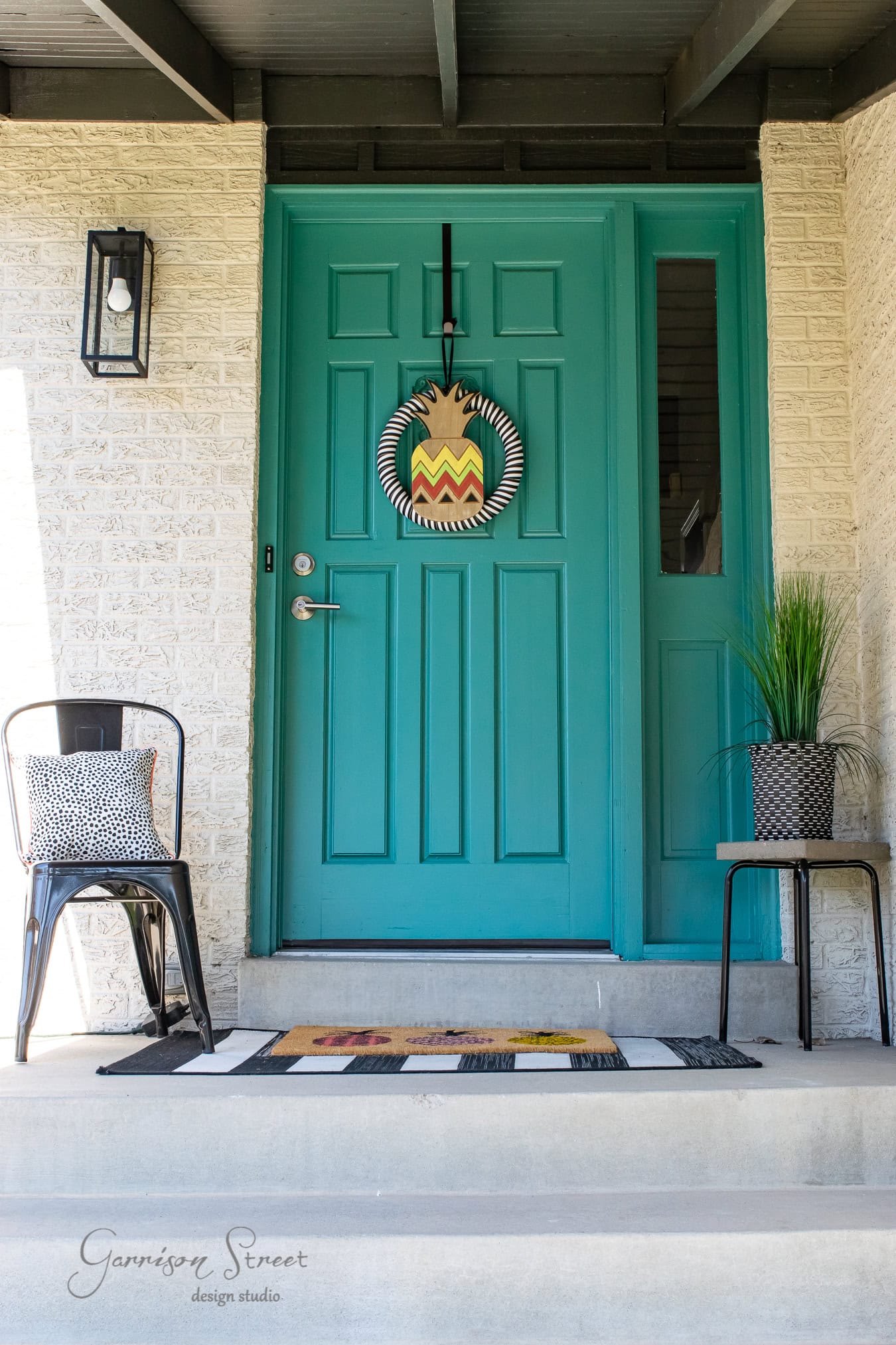 A bright teal front door, set in a classic brick facade, is adorned with a colorful wreath. A black chair with a patterned cushion sits on the porch next to a small table holding a potted plant. A striped doormat lies in front of the door, boasting "Garrison Street design studio".