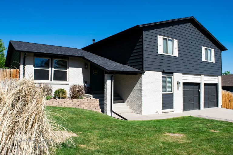 A modern house with a chic black and white exterior, featuring two stories and a spacious two-car garage. The front yard is landscaped with grass and dry plants against a clear blue sky, highlighting its sleek design.