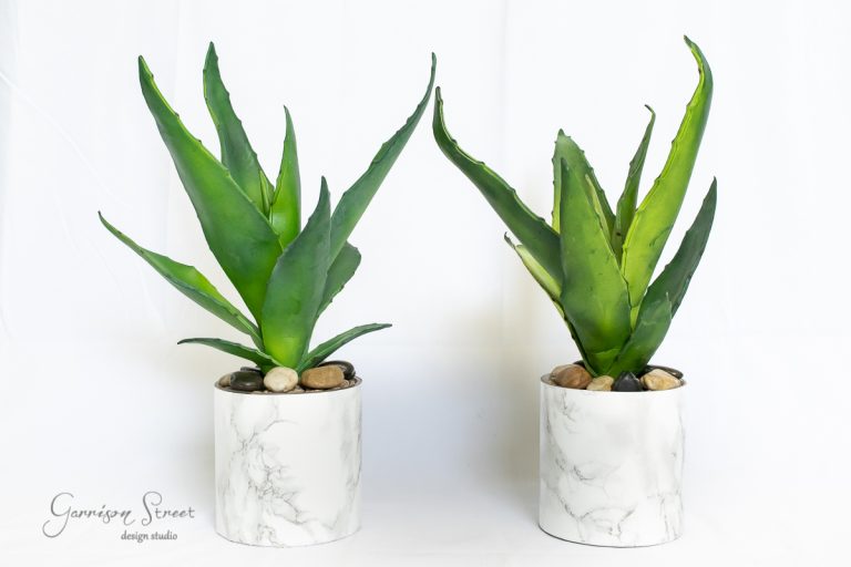 Two artificial agave plants in white marble-patterned pots with pebbles. The plants have long, pointed green leaves and are placed against a white background.