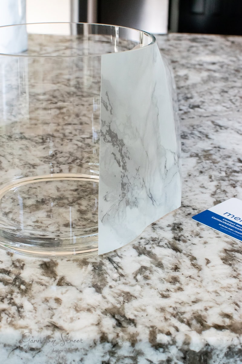 A clear glass container with a piece of marbled adhesive paper partially attached sits on a speckled granite countertop, resembling a sleek planter. A blue and white instruction card lies nearby.